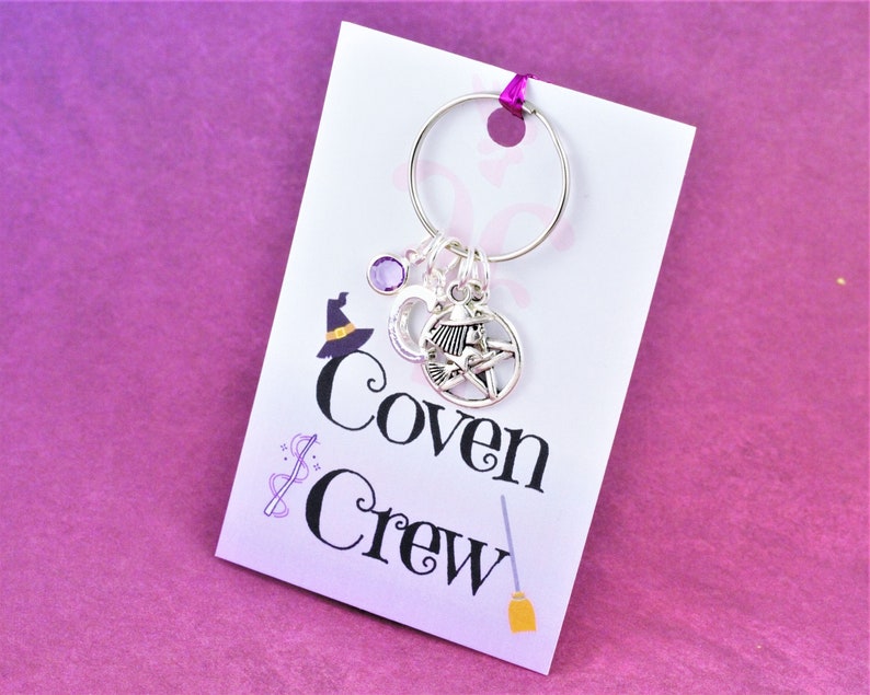 Puede incluir: Una tarjeta blanca con el texto "Coven Crew" y una peque&ntilde;a escoba impresa en ella. Un llavero plateado con una piedra preciosa p&uacute;rpura, un amuleto de luna creciente y un amuleto de pentagrama est&aacute; unido a la tarjeta.