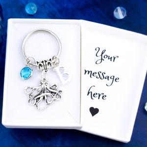 May include: A silver octopus keychain with a blue crystal charm and a silver letter B charm. The keychain is in a white gift box with the text "Your message here" and a black heart.