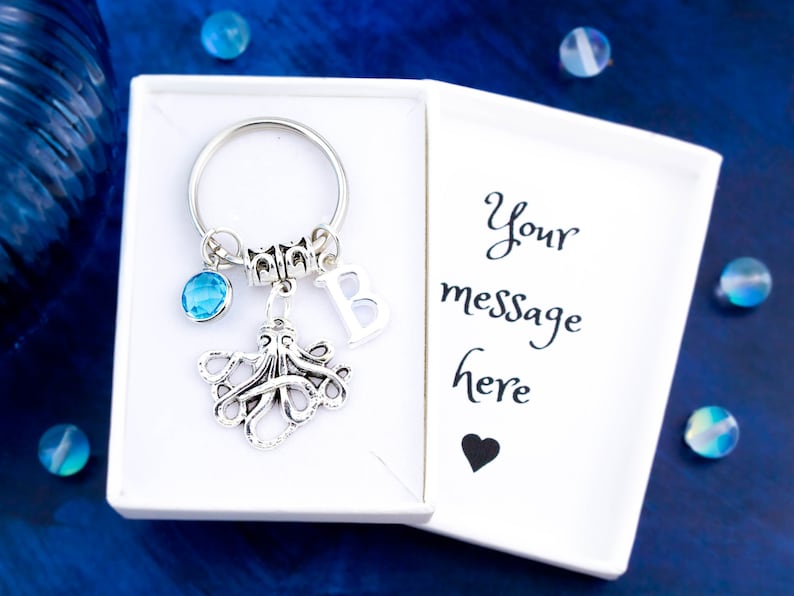 May include: A silver octopus keychain with a blue crystal charm and a silver letter B charm. The keychain is in a white gift box with the text "Your message here" and a black heart.
