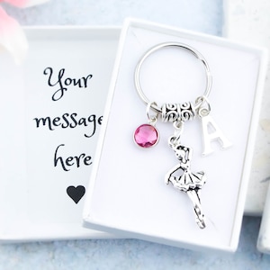 May include: A silver ballerina keychain with a pink rhinestone, a silver letter A charm, and a silver key ring. The keychain is in a white gift box with the text "Your message here" on the lid.