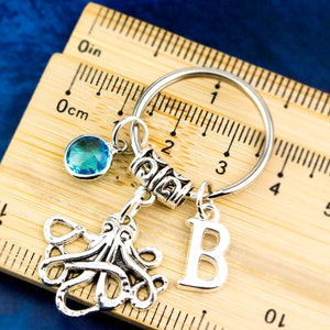 May include: Silver keychain with a blue crystal charm, an octopus charm, and a letter 'B' charm. The keychain is on a wooden ruler with inch and centimeter markings.