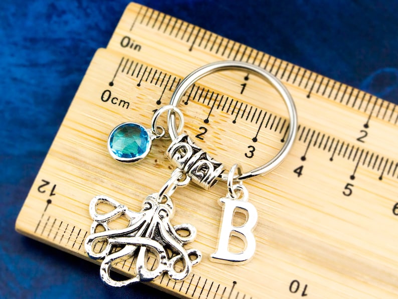 May include: Silver keychain with a blue crystal charm, an octopus charm, and a letter 'B' charm. The keychain is on a wooden ruler with inch and centimeter markings.