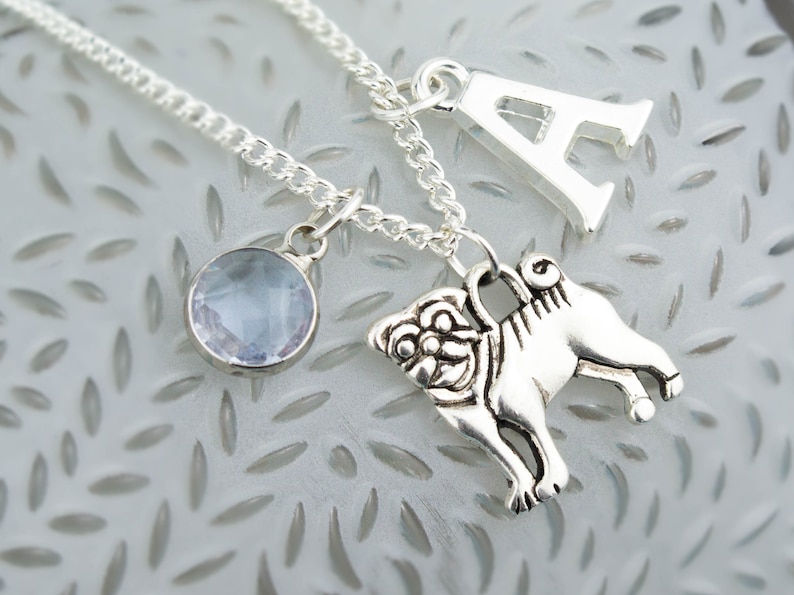 May include: Silver necklace with a light blue gemstone, a silver pug charm, and a silver letter A charm.