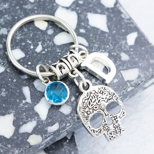 May include: Silver keychain with a blue crystal charm, a silver skull charm with floral details, and a silver charm with the letter "D".