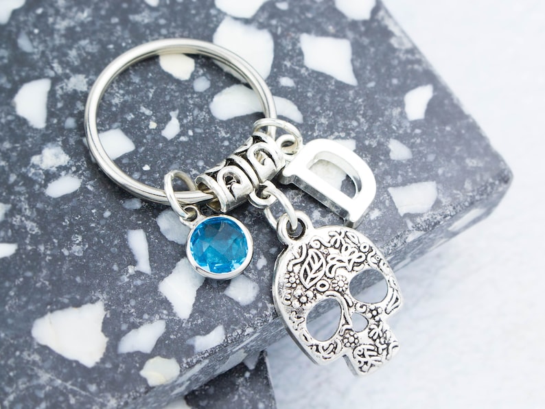 May include: Silver keychain with a blue crystal charm, a silver skull charm with floral details, and a silver charm with the letter "D".