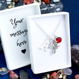 May include: A silver necklace with a red crystal charm, a silver letter A charm, and a silver dragon charm. The necklace is in a white gift box with a blank card that says "Your message here".