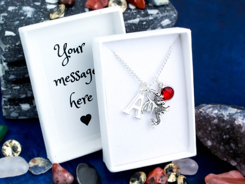 May include: A silver necklace with a red crystal charm, a silver letter A charm, and a silver dragon charm. The necklace is in a white gift box with a blank card that says "Your message here".