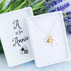 Puede incluir: Una caja de regalo blanca con un collar de plata en su interior. El collar tiene un dije de abeja y un dije con la letra A. La caja tiene el texto "A is for Annie" y una ilustración de una abeja.