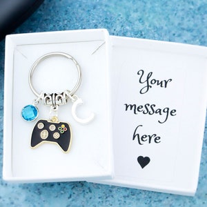 May include: A silver keyring featuring a black video game controller charm, a blue gemstone, and a letter "C" charm. The keyring is presented in a white gift box with the words "Your message here".
