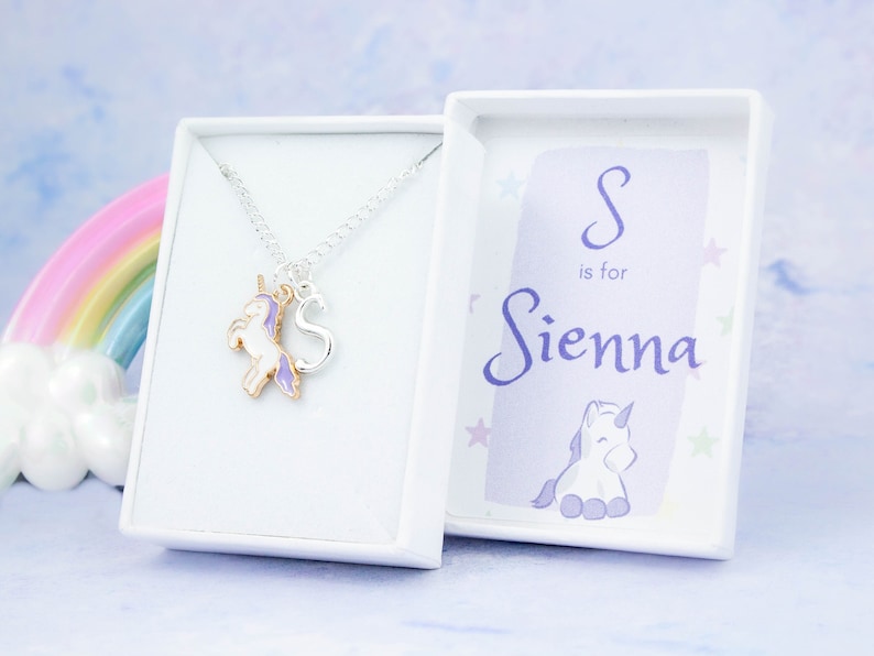 Op de afbeelding: Een zilveren ketting met een paarse en witte eenhoornbedel en een zilveren letter 'S'-bedel. De ketting wordt geleverd in een witte doos met een paarse en witte eenhoorn en de tekst 'S is voor Sienna'.