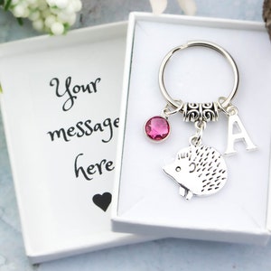 May include: A silver hedgehog keychain with a pink crystal charm, a silver letter A charm, and a silver key ring. The keychain is in a white gift box with the text "Your message here" on the lid.