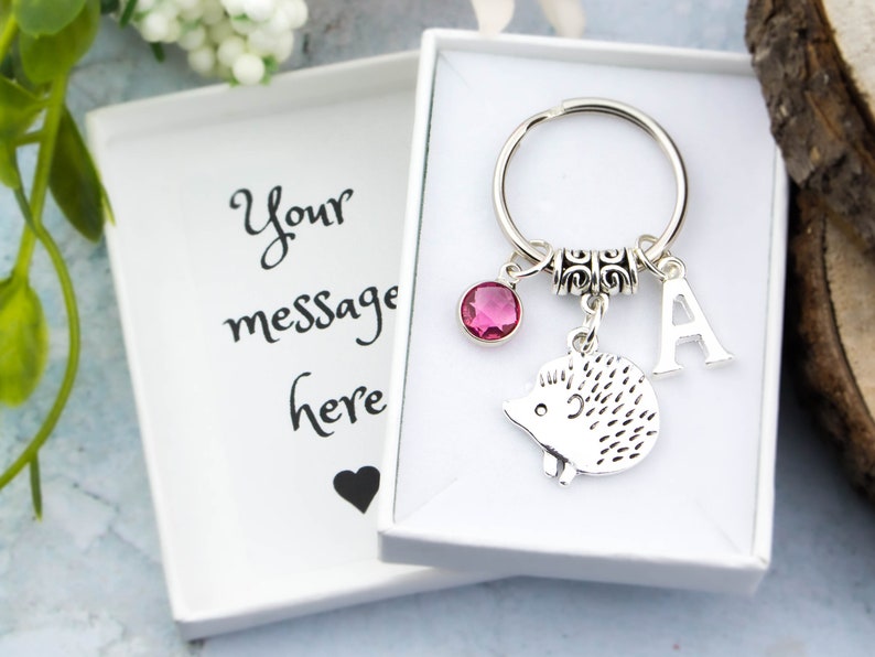 May include: A silver hedgehog keychain with a pink crystal charm, a silver letter A charm, and a silver key ring. The keychain is in a white gift box with the text "Your message here" on the lid.