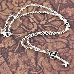 May include: A silver key necklace with a detailed key charm. The key has a decorative design and a small loop at the top. The chain is a delicate silver chain.
