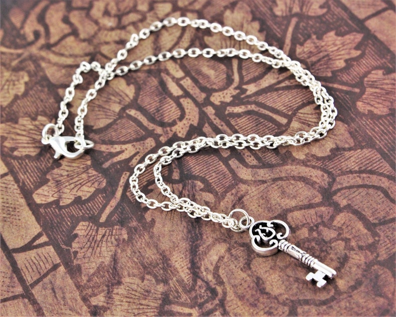 May include: A silver key necklace with a detailed key charm. The key has a decorative design and a small loop at the top. The chain is a delicate silver chain.