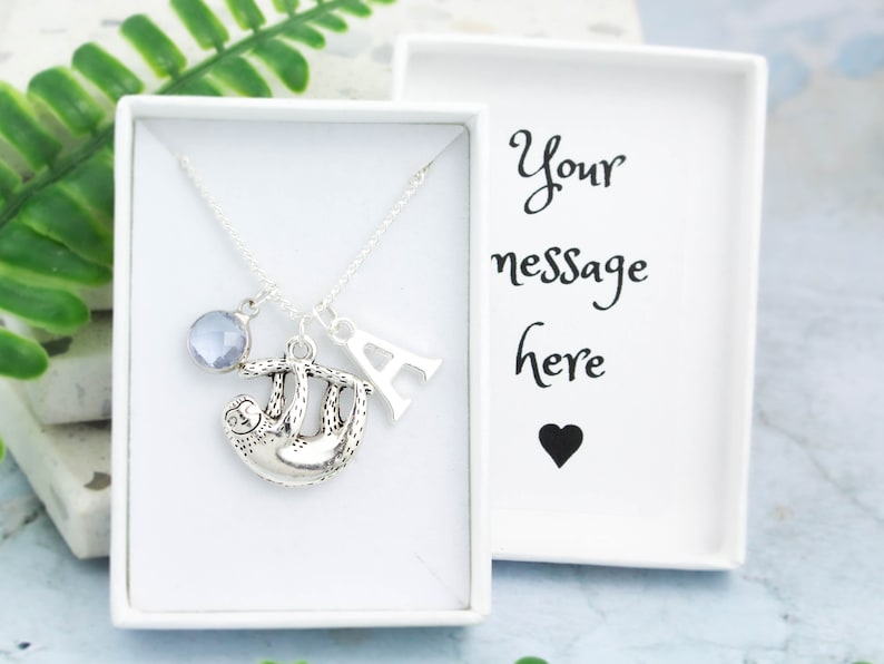 May include: A silver necklace with a sloth charm, a blue crystal, and the letter A. The necklace is in a white gift box with the text "Your message here" and a black heart.