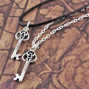 May include: Two silver key necklaces with intricate designs. One necklace has a black cord and the other has a silver chain.