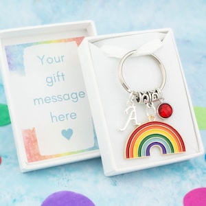 May include: A rainbow keyring with a red gem, a silver letter A charm, and a silver ring. The keyring is in a white gift box with a white ribbon. The box has a card that says "Your gift message here" with a heart.