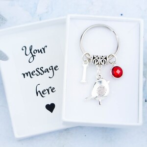 May include: A silver keyring with a bird charm, a red gem, and the letter "I" inside a white gift box. The box lid has the text "Your message here" and a black heart.