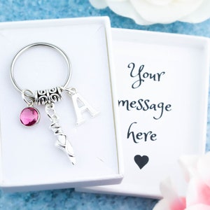 Puede incluir: Un llavero plateado con un dije de cristal rosa, un dije de bailarina plateada y un dije de letra A plateada. El llavero está en una caja de regalo blanca con el texto "Tu mensaje aquí" y un corazón negro.