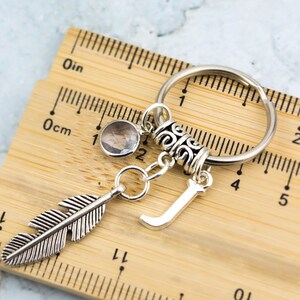 Puede incluir: Llavero de plata con un dije de pluma, un dije de cristal transparente, un dije de letra J de plata y un anillo decorativo de plata. El llavero se mide contra una regla de madera.