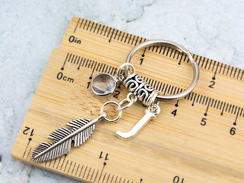Puede incluir: Llavero de plata con un dije de pluma, un dije de cristal transparente, un dije de letra J de plata y un anillo decorativo de plata. El llavero se mide contra una regla de madera.