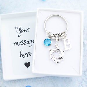 May include: A silver keyring with a blue crystal charm, a dolphin charm, and a letter "B" charm. The keyring is in a white box with the text "Your message here" on the inside.
