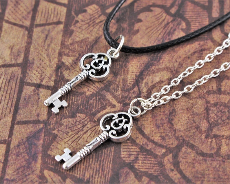 May include: Two silver key necklaces, one on a black cord and the other on a silver chain. The keys have ornate designs and are a popular choice for couples.