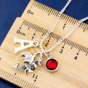 May include: Silver necklace with a dragon charm, a letter A charm, and a red gemstone charm. The necklace is measured against a ruler.