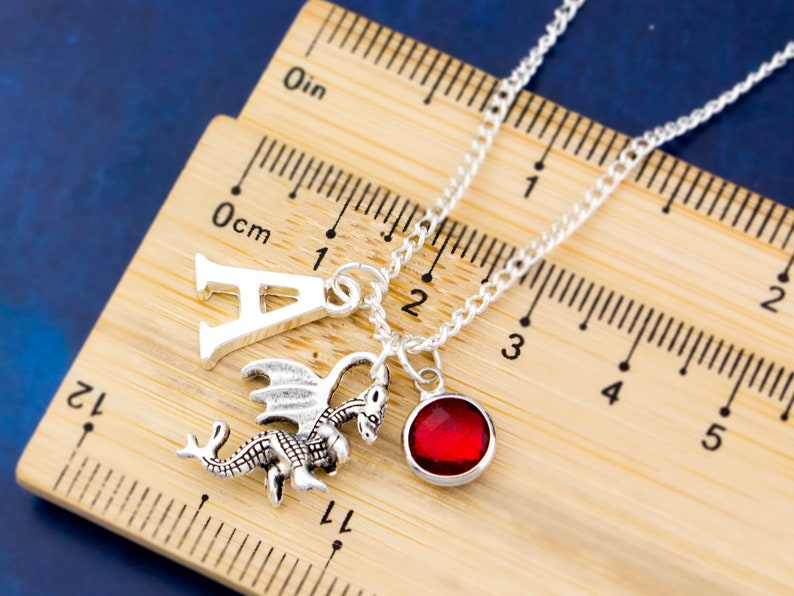May include: Silver necklace with a dragon charm, a letter A charm, and a red gemstone charm. The necklace is measured against a ruler.