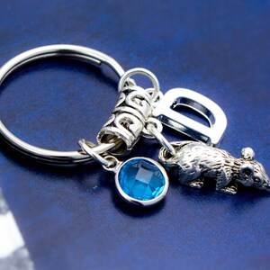 Puede incluir: Llavero de plata con un dije de cristal azul, un dije de letra D de plata y un dije de rata de plata. El llavero est&aacute; unido a un anillo de llave de plata.