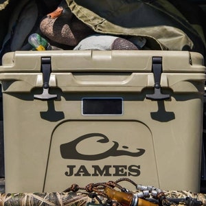 May include: A tan cooler with a black duck logo and the name "JAMES" printed on the side. The cooler is in the back of a pickup truck with other hunting gear.