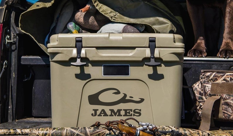 May include: A tan cooler with a black duck logo and the name "JAMES" printed on the side. The cooler is in the back of a pickup truck with other hunting gear.