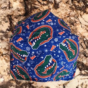 May include: Blue fabric item with a repeating pattern of green alligators, orange letter "F" logos, and white floral accents. The item is likely a face covering or hat.