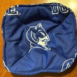 May include: A square, blue bowl cozy featuring the Duke Blue Devils logo in white. The logo is a stylized image of a devil. The bowl cozy is made of quilted fabric and has white text around the edges.
