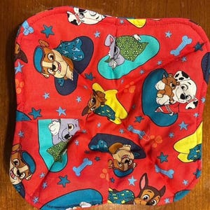 May include: A red, square-shaped quilted pot holder featuring a repeating pattern of cartoon puppies, blue stars, and bone shapes. The puppies are in various poses on blue and yellow backgrounds.