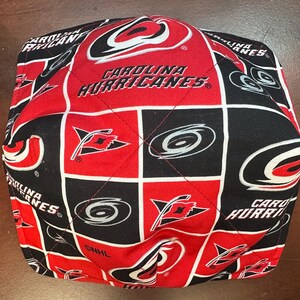 May include: A quilted bowl cozy featuring a repeating pattern of the Carolina Hurricanes logo and team name in red, black, and white. The design includes the team's logo and the words "CAROLINA HURRICANES".