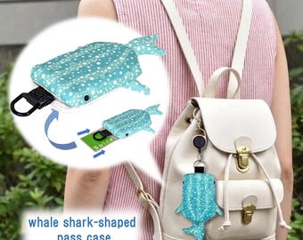 May include: A teal whale shark-shaped pass case with white polka dots, featuring a retractable reel and a black clip. The case is shown attached to a white backpack and includes a diagram demonstrating how to insert a card into the holder.