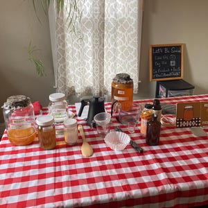 May include: A kombucha brewing setup featuring large glass jars with cloth covers, a black kettle, a wooden spoon, glass measuring cups, a funnel, paper filters, and amber glass bottles. A small chalkboard sign reads Let's Be Social Like Us Tag Us.