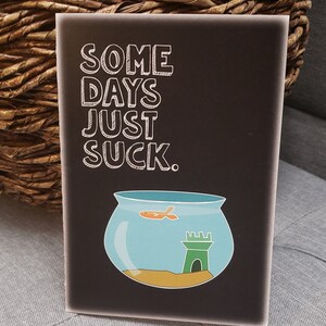 Pode incluir: Um cartão preto com texto branco que diz "Some days just suck." Uma tigela de peixe de desenho animado com um peixe dourado e um castelo verde está abaixo do texto.