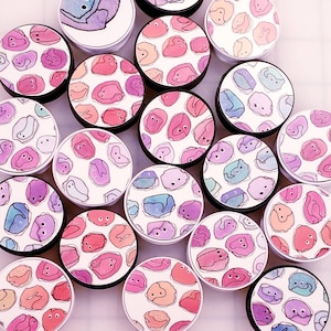 May include: A collection of colorful round stickers with various whimsical designs featuring cartoon faces and abstract patterns. The stickers are arranged in a circular pattern on a white background.