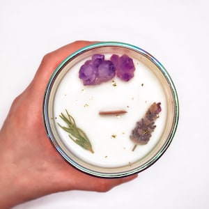 May include: A white candle with amethyst crystals, lavender, and rosemary embedded in the wax. The candle is in a clear glass jar with an iridescent finish.