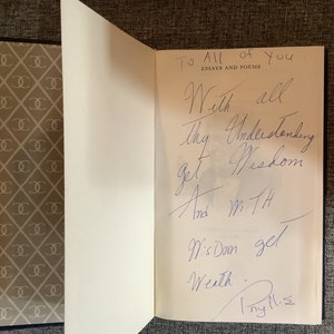 May include: A white book with a blue cover. The book is open to a page with a handwritten inscription that reads "To All of You. Essays and Poems. With all Thy Understanding get Wisdom And with Wisdom get Wealth. Thy M.S."