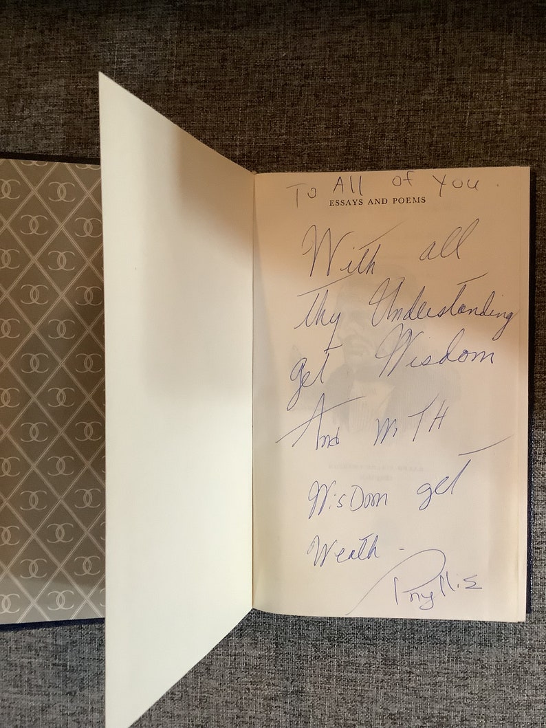 May include: A white book with a blue cover. The book is open to a page with a handwritten inscription that reads "To All of You. Essays and Poems. With all Thy Understanding get Wisdom And with Wisdom get Wealth. Thy M.S."