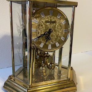 May include: A gold-toned, glass-enclosed clock with a decorative floral design etched on the glass. The clock face is gold with black Roman numerals and ornate detailing. The clock has a pendulum and weights.