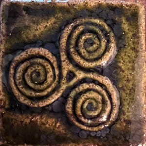 Op de afbeelding: Vierkante keramische tegel met een verhoogd ontwerp van drie spiralen. De spiralen zijn goudbruin, tegen een gevlekte achtergrond van groen en bruin. De tegel heeft een glanzende afwerking.