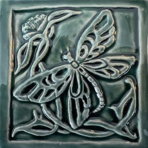 May include: Square ceramic tile in teal green, featuring a raised relief design of a dragonfly and floral elements. The dragonfly has detailed wings, surrounded by stylized leaves and a flower, creating a nature-inspired motif.