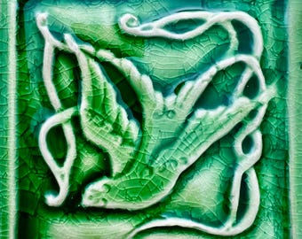 May include: A green ceramic tile with a white raised design of a bird in flight. The tile has a crackled glaze.