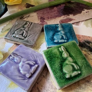 Può includere: Quattro piastrelle in ceramica con un design rialzato di un coniglio in diversi colori: verde, blu, viola e grigio.