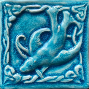 Puede incluir: Un azulejo de cerámica azul con un diseño blanco de un animal estilizado, posiblemente un gato o un pájaro, con un borde floral.