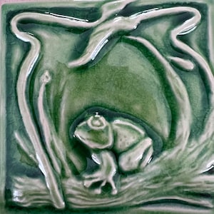 以下が含まれることがあります： カエルが睡蓮の葉の上にいるレリーフデザインが施された、正方形の緑色のセラミックタイル。カエルは明るい緑色で、背景は濃い緑色です。タイルは光沢のある仕上がりです。